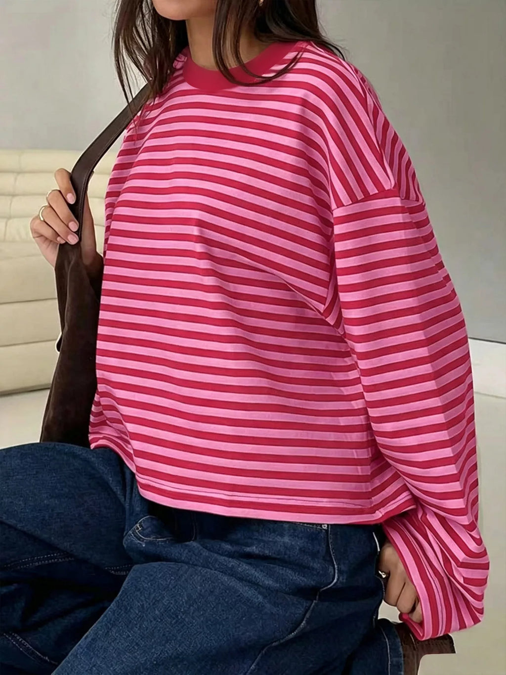 Side profile of seated model in pink and deep red striped tee tucked into dark blue jeans holding brown suede bag strap