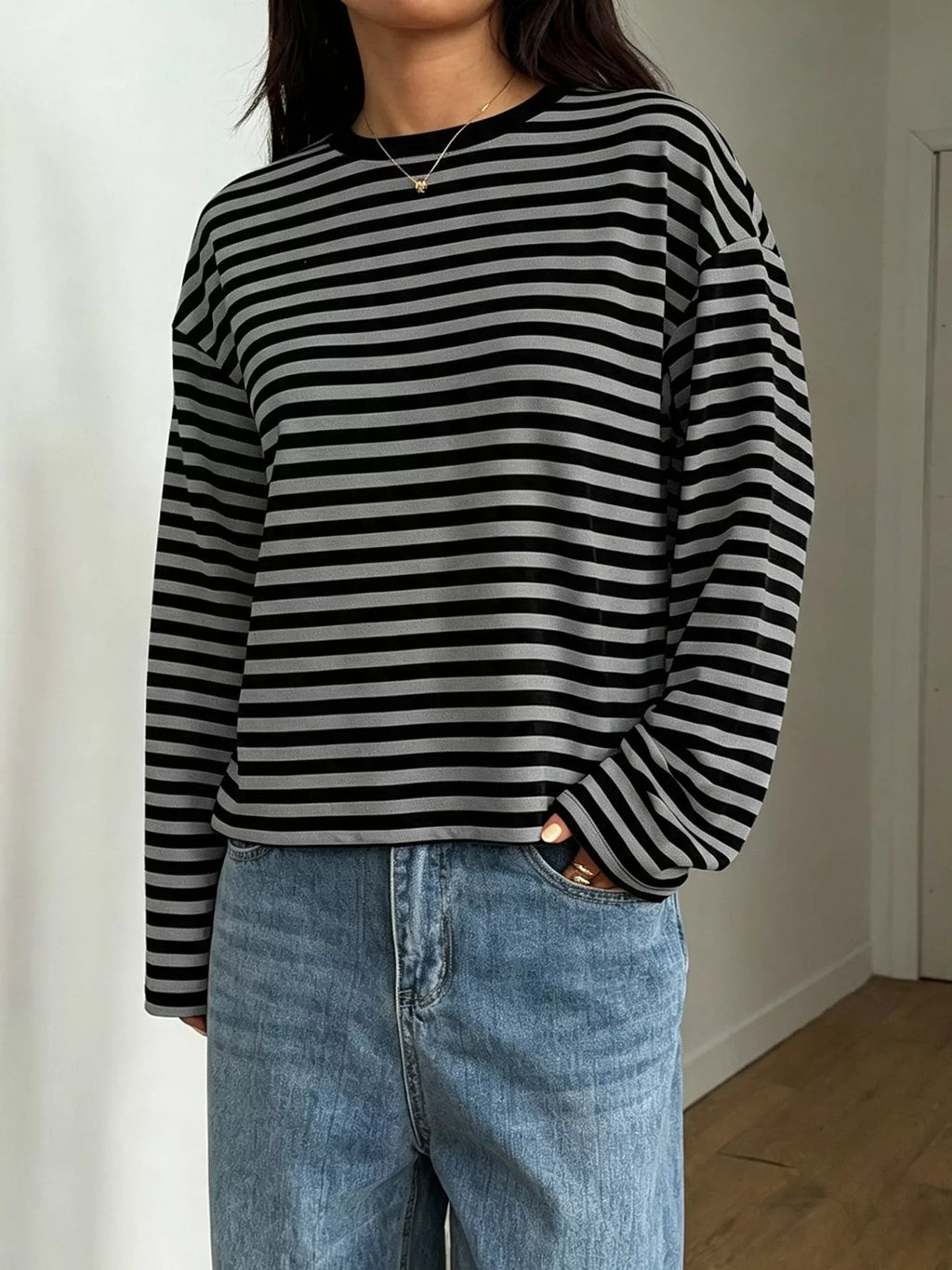 Model in black and grey striped long sleeve tee with relaxed fit, blue jeans, and gold necklace against white wall