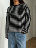 Model in black and grey striped long sleeve tee with relaxed fit, blue jeans, and gold necklace against white wall