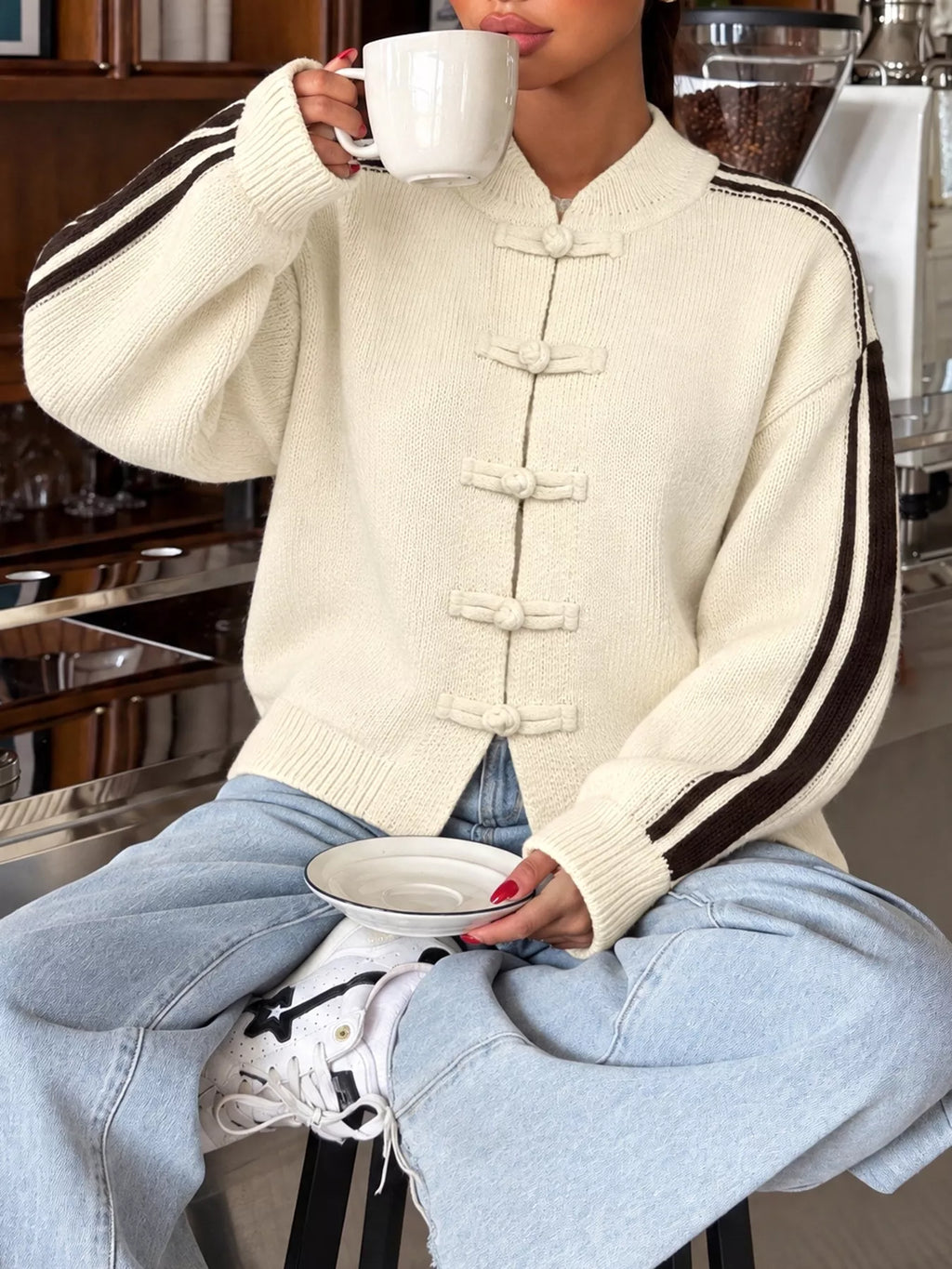 Modelin creamfarbener Strickjacke mit Chinesischen Knotenknöpfen und braunen Seitenstreifen, die eine Tasse hält
