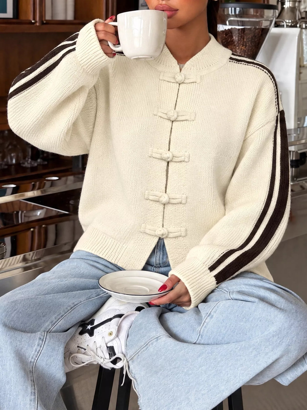 Modelin creamfarbener Strickjacke mit Chinesischen Knotenknöpfen und braunen Seitenstreifen, die eine Tasse hält