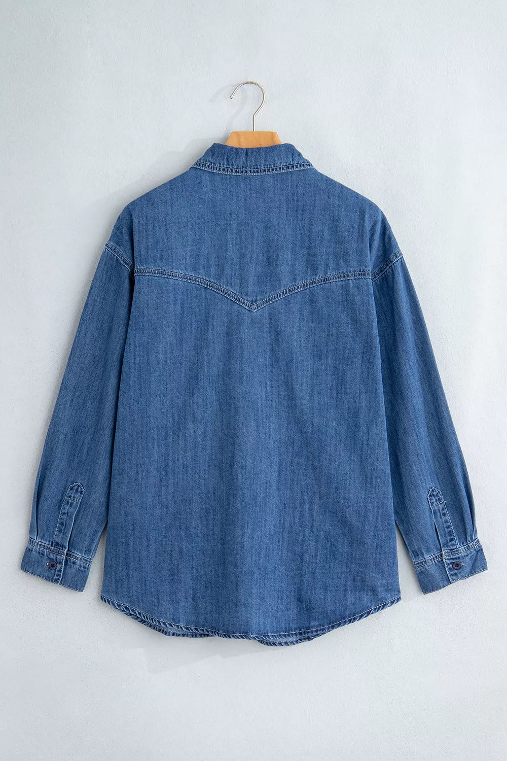 Vista trasera camisa vaquera en percha: costura arco continua espalda superior, puños ajustables y fondo redondeado.