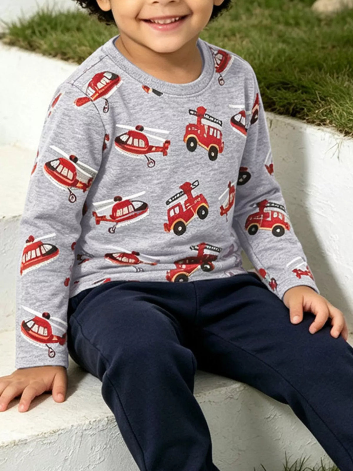 Boy sitting on concrete ledge wearing grey vehicle graphic t-shirt with navy trousers outdoors
