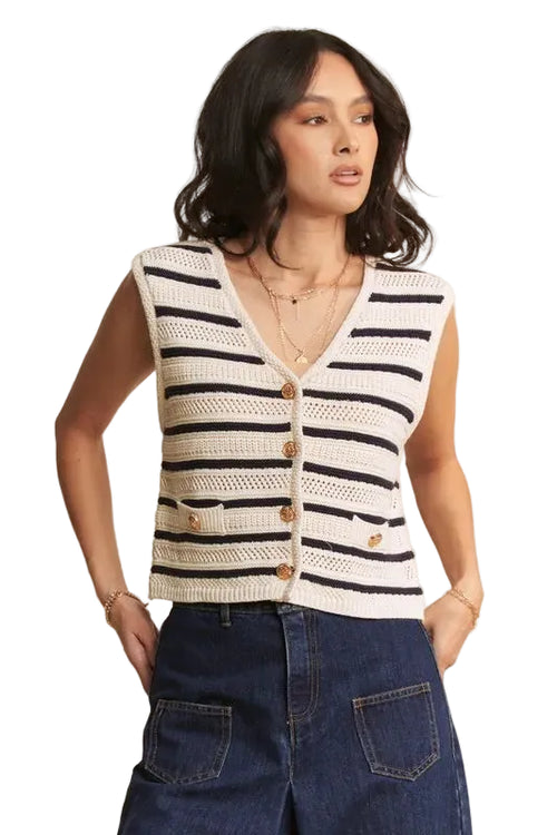 Model in navy cream striped knit vest with openwork mesh, V-neck, gold buttons, and layered necklaces on beige background