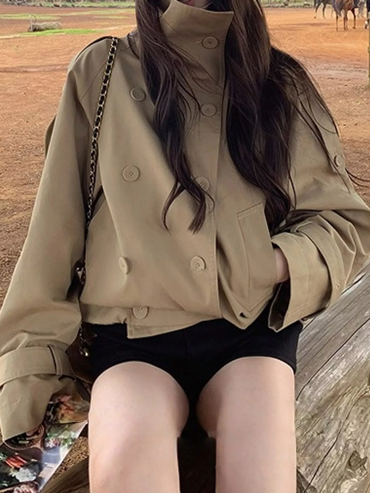 Sitzendes Model im beigen Crop-Trench mit hohem Kragen und funktionalem Knopfband an den Ärmeln
