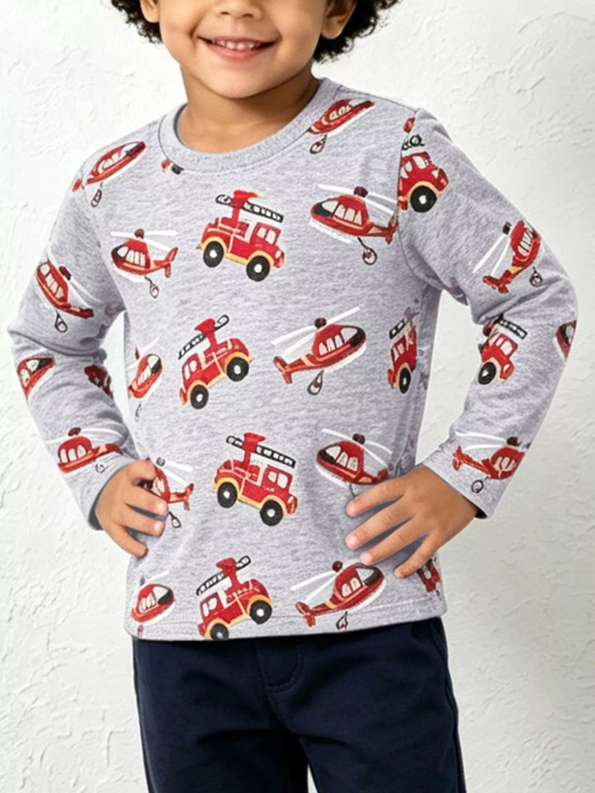 Young boy in heather grey long sleeve crew neck tee with red fire truck and helicopter print against white wall