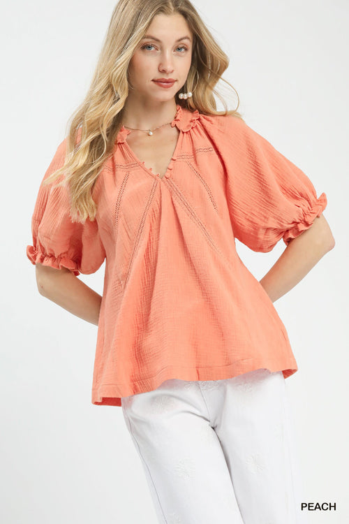 Model in peach cotton gauze blouse with V-neck ruffle trim, lace inserts, and puff sleeves paired with white pants