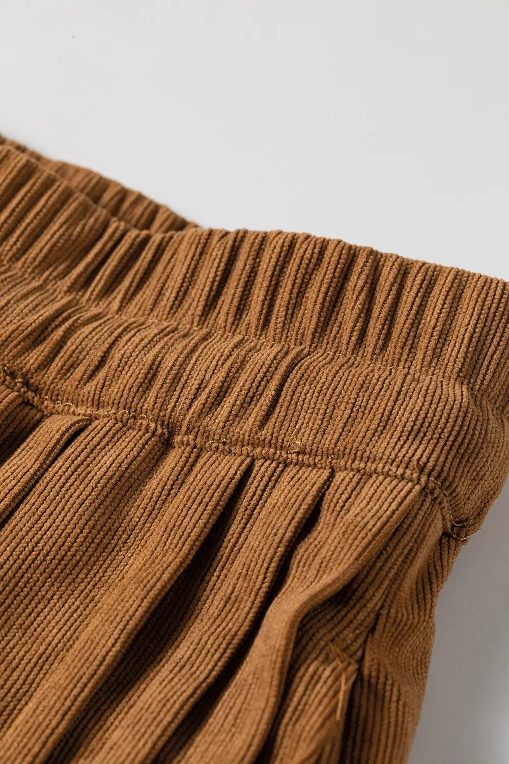 Primer plano de cintura elástica recogida en pantalones de pana camel, con costuras verticales que conectan con los pliegues