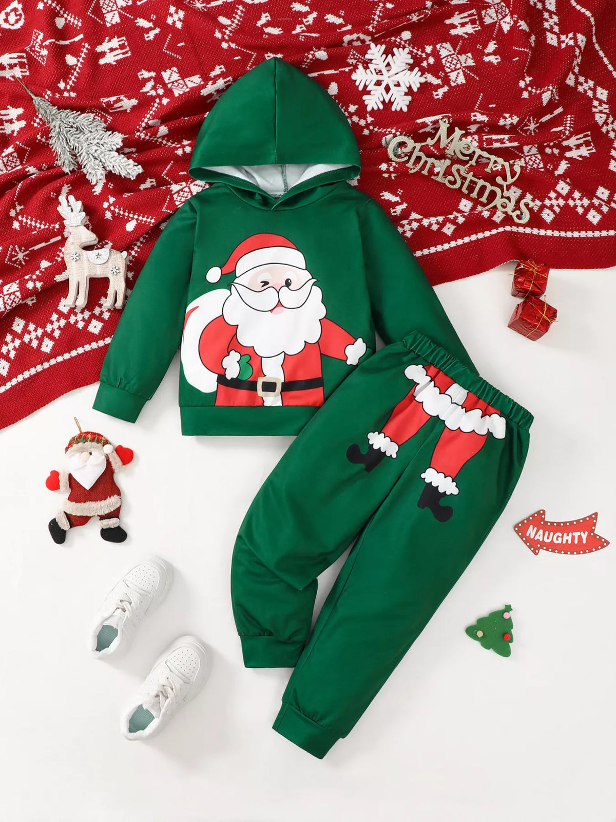 Green Santa graphic hoodie and pants set flat lay with red blanket, white sneakers, wooden reindeer, and Christmas ornaments