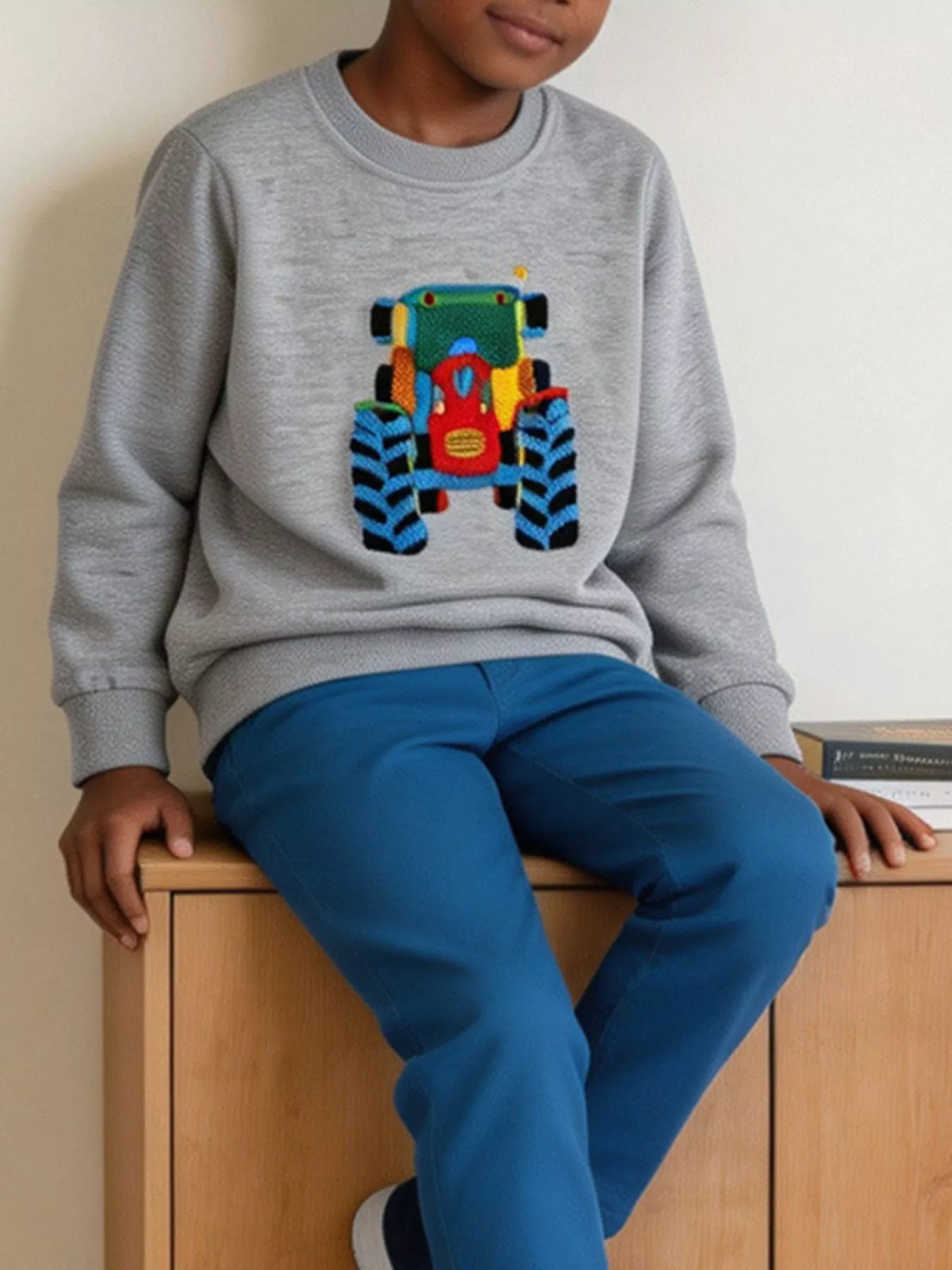 Kind sitzt auf Holzfläche und trägt graues Rundhals-Sweatshirt mit zentralem Fahrzeug-Stickerei-Design