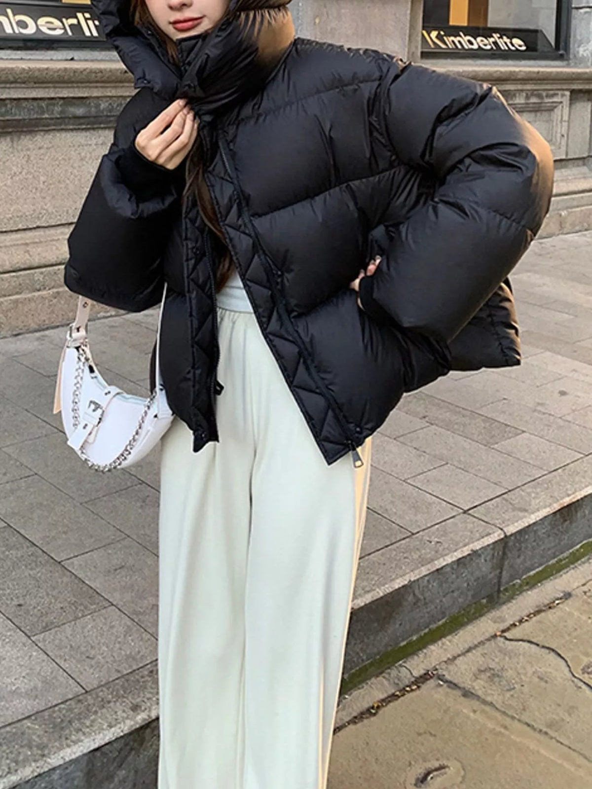 Woman wearing black quilted puffer with hood up, carrying white crescent bag for street style look
