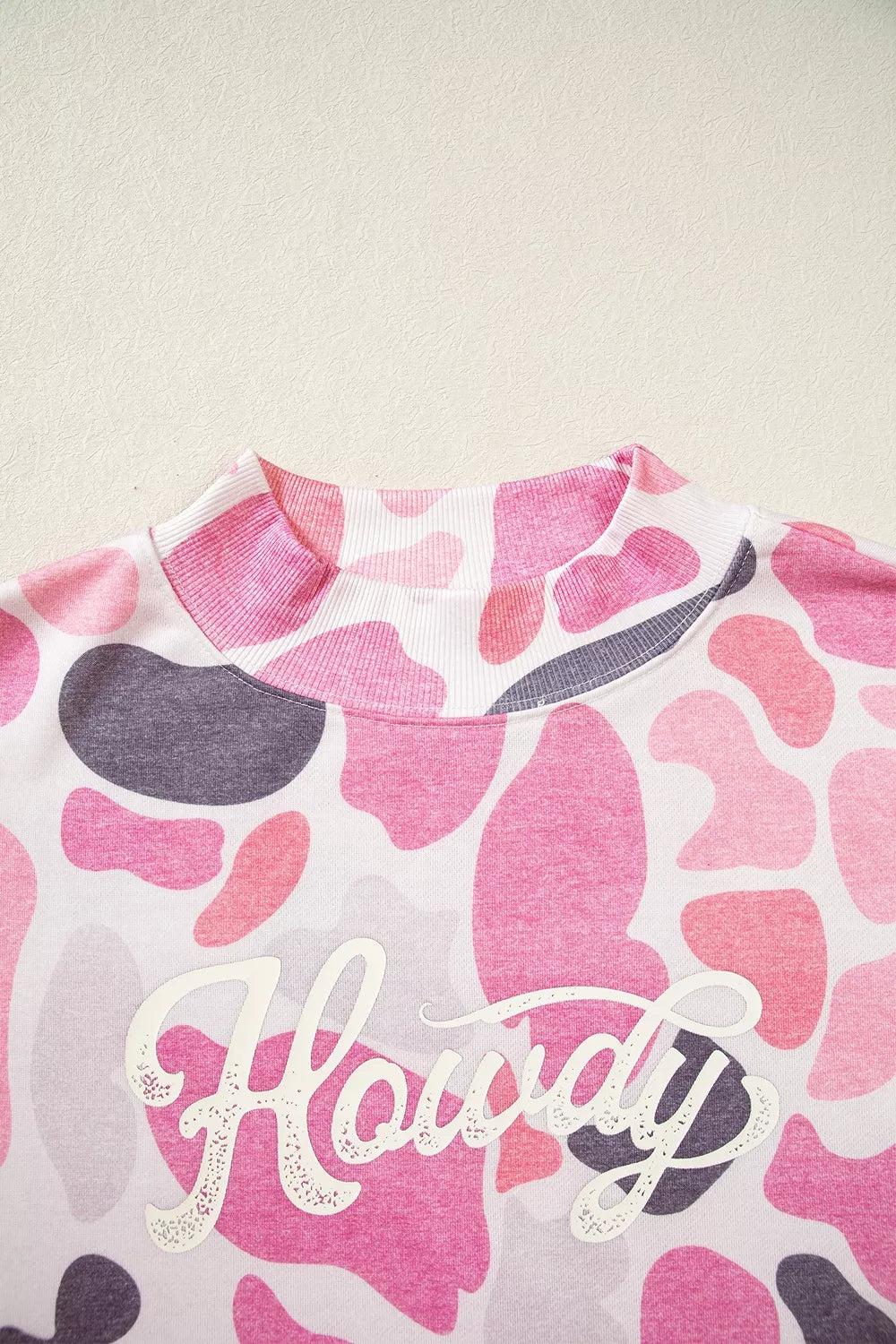 White cursive Howdy text graphic in distressed font overlaid on pink grey camo print