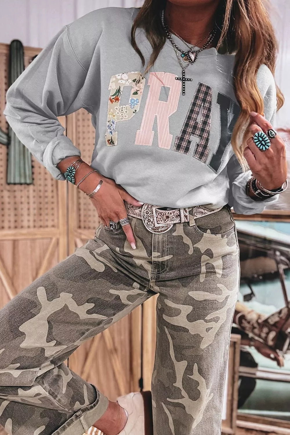 Front view of grey patchwork sweatshirt paired with high-waisted camo denim jeans and ornate western belt buckle. Model wears