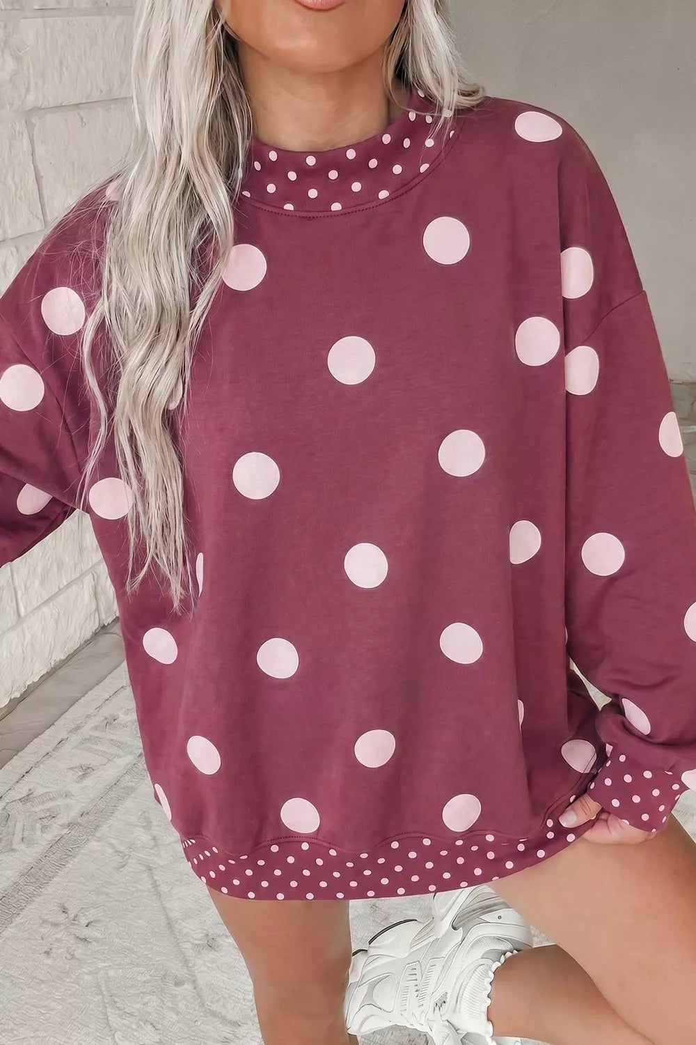 Model in oversized maroon polka dot tunic sweatshirt with white sneakers against white brick wall