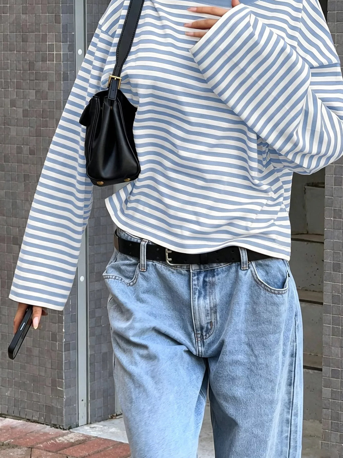T-shirt maniche lunghe a righe blu e bianche inserita nei jeans acid wash con cintura nera e borsa crossbody in ambiente urbano