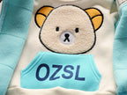 Macro view of cream sweatshirt chest with high-pile beige bear applique and blue OZSL printed pocket