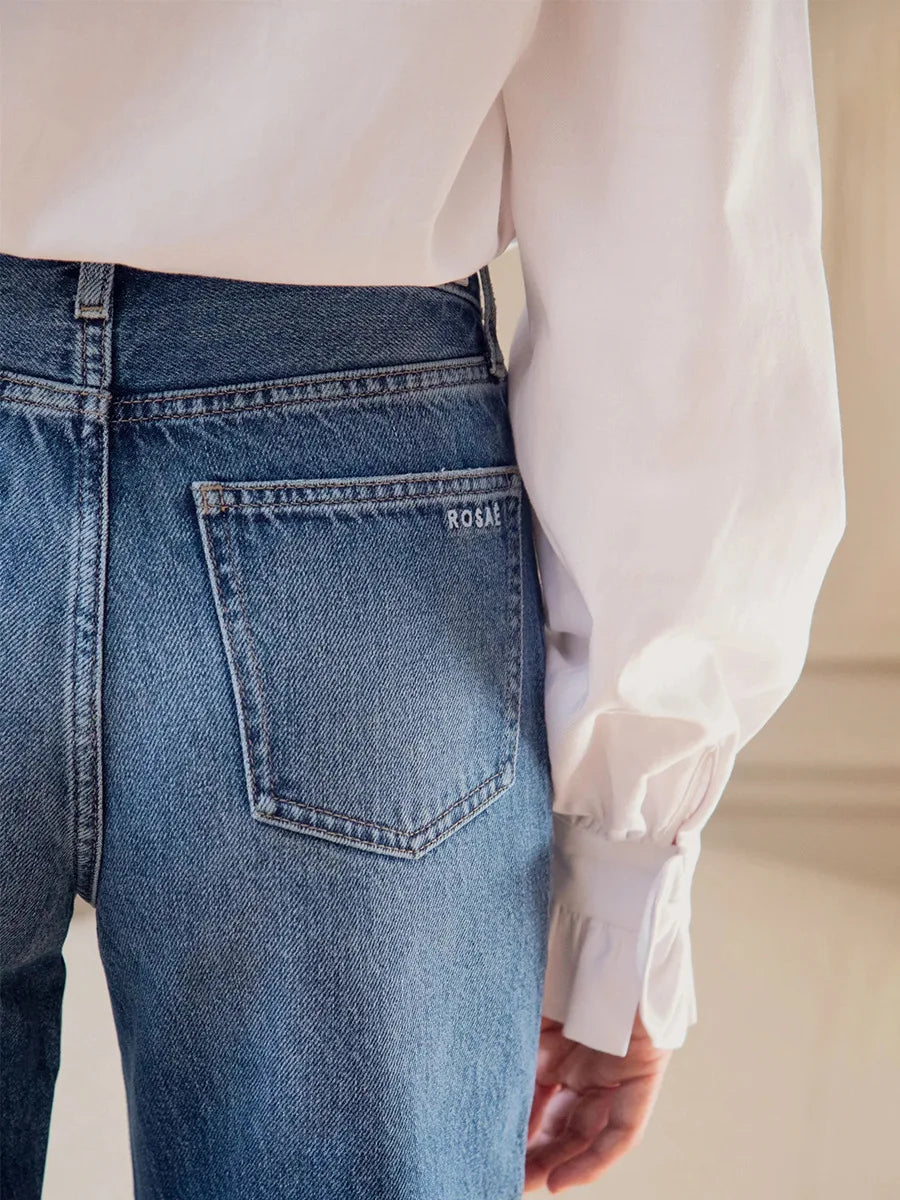 Detailaufnahme: Blaue Jeans mit 'ROSAI'-Stickerei kombiniert mit weißer Bluse und gerafften Rüschenärmeln