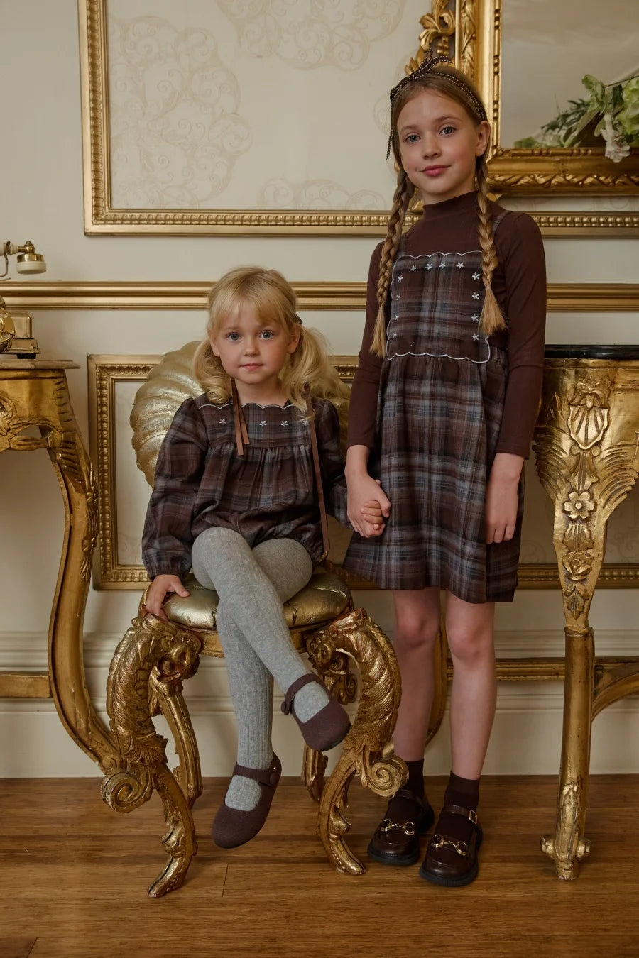 Duas meninas em vestidos de alça com detalhes bordados brancos, posando perto de mobiliário dourado.