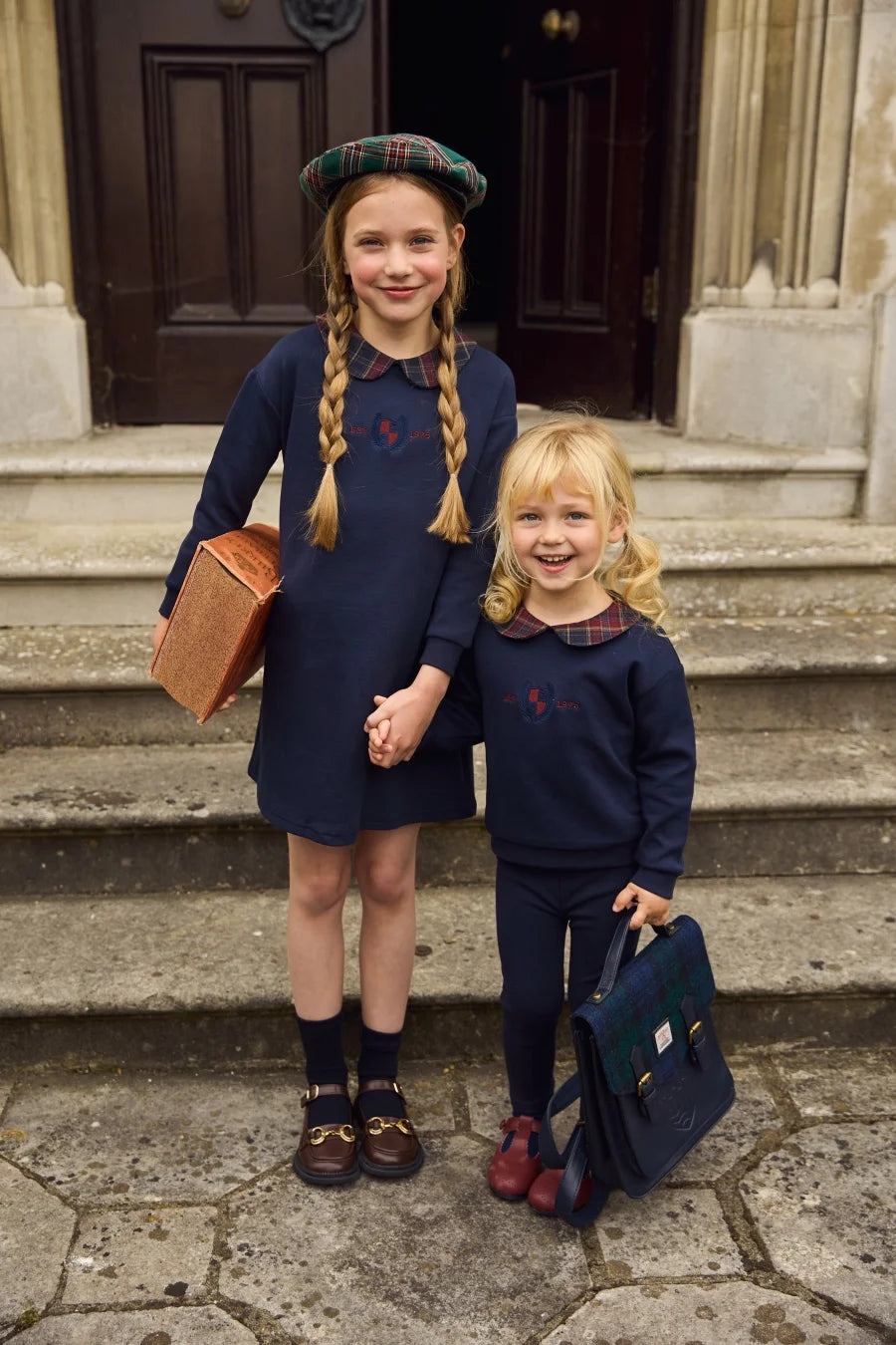 Duas meninas em conjuntos azuis-marinho combinados com colarinhos tartan e bordado no peito, usando boinas verdes e mocassins.