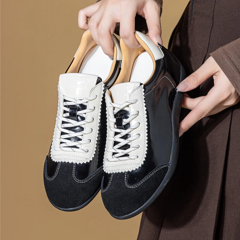 Primer plano de zapatillas negras sujetadas por manos mostrando parte superior brillante, puntera de ante y panel de cordón cosido en zigzag