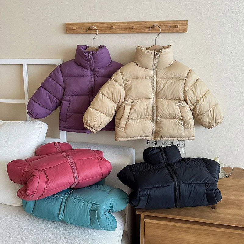 Five solid color kids winter coats in purple, beige, pink, teal, and black displayed on a rack and furniture showing