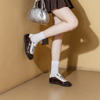 Foto de moda de cuerpo entero de modelo con zapatillas marrones y falda plisada sosteniendo bolso plateado contra pared amarilla
