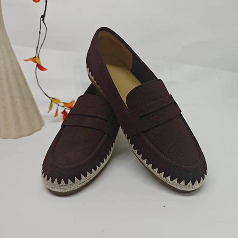 Mocassini slip-on marrone scuro con punta tonda e cuciture bianche seghettate accanto a foglie arancioni