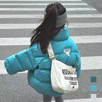 Child in bright turquoise quilted jacket crossing street with grey headband and white canvas shoulder bag.