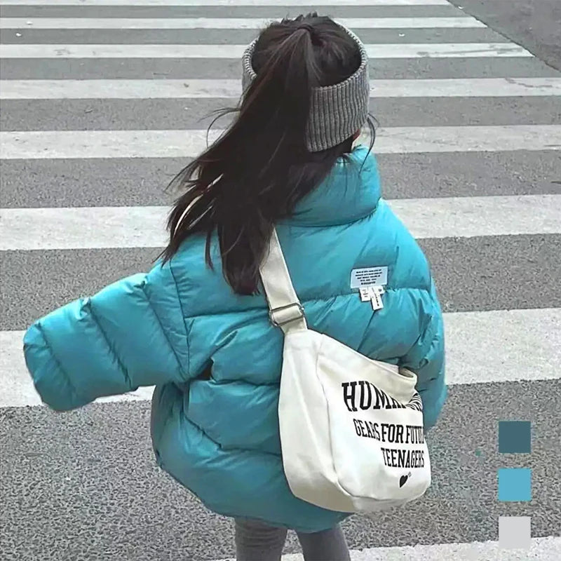 Child in bright turquoise quilted jacket crossing street with grey headband and white canvas shoulder bag.