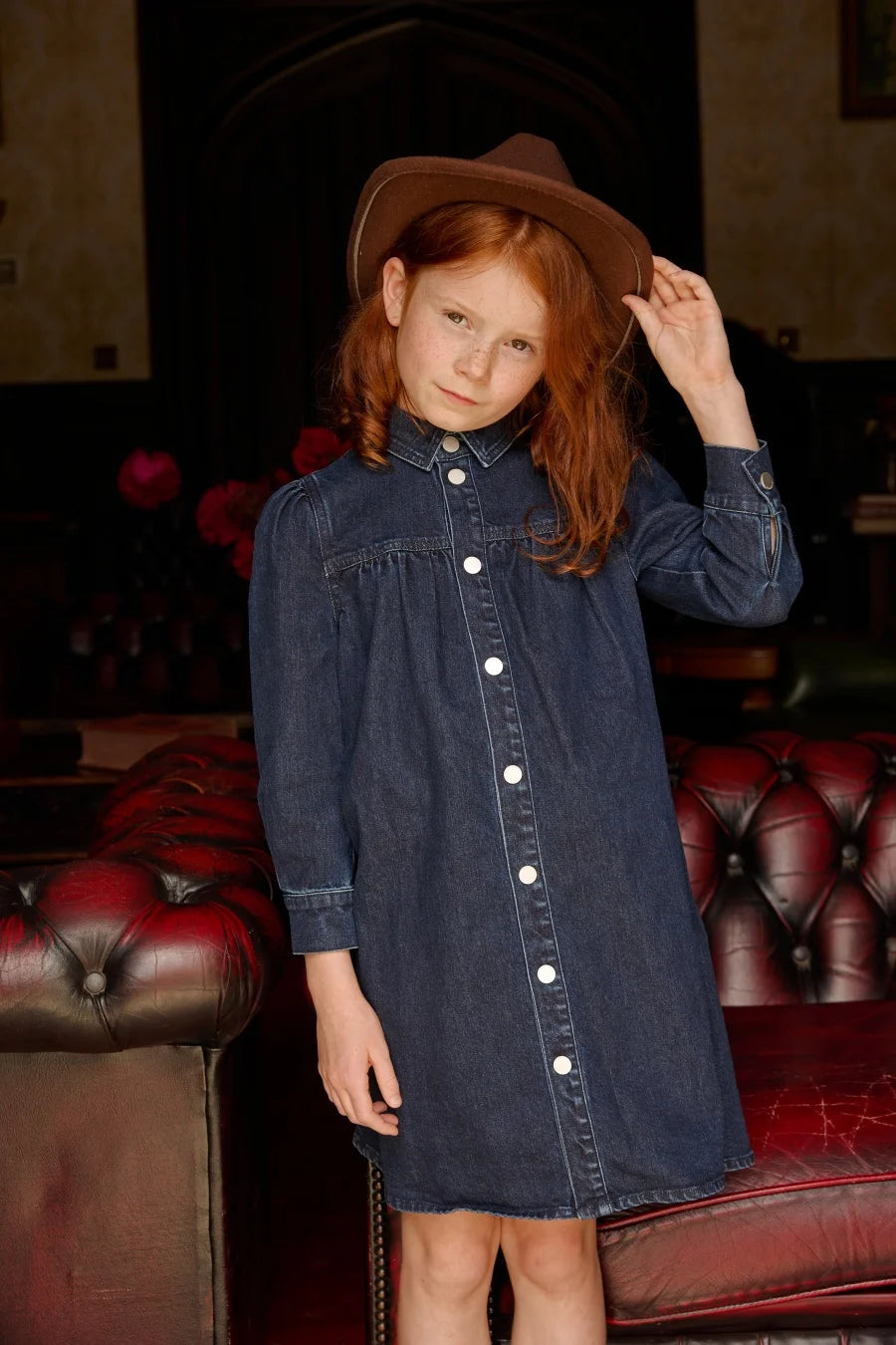 Menina ruiva a experimentar vestido-camisa de ganga azul-escuro com pregas e botões brancos junto a um sofá vermelho.