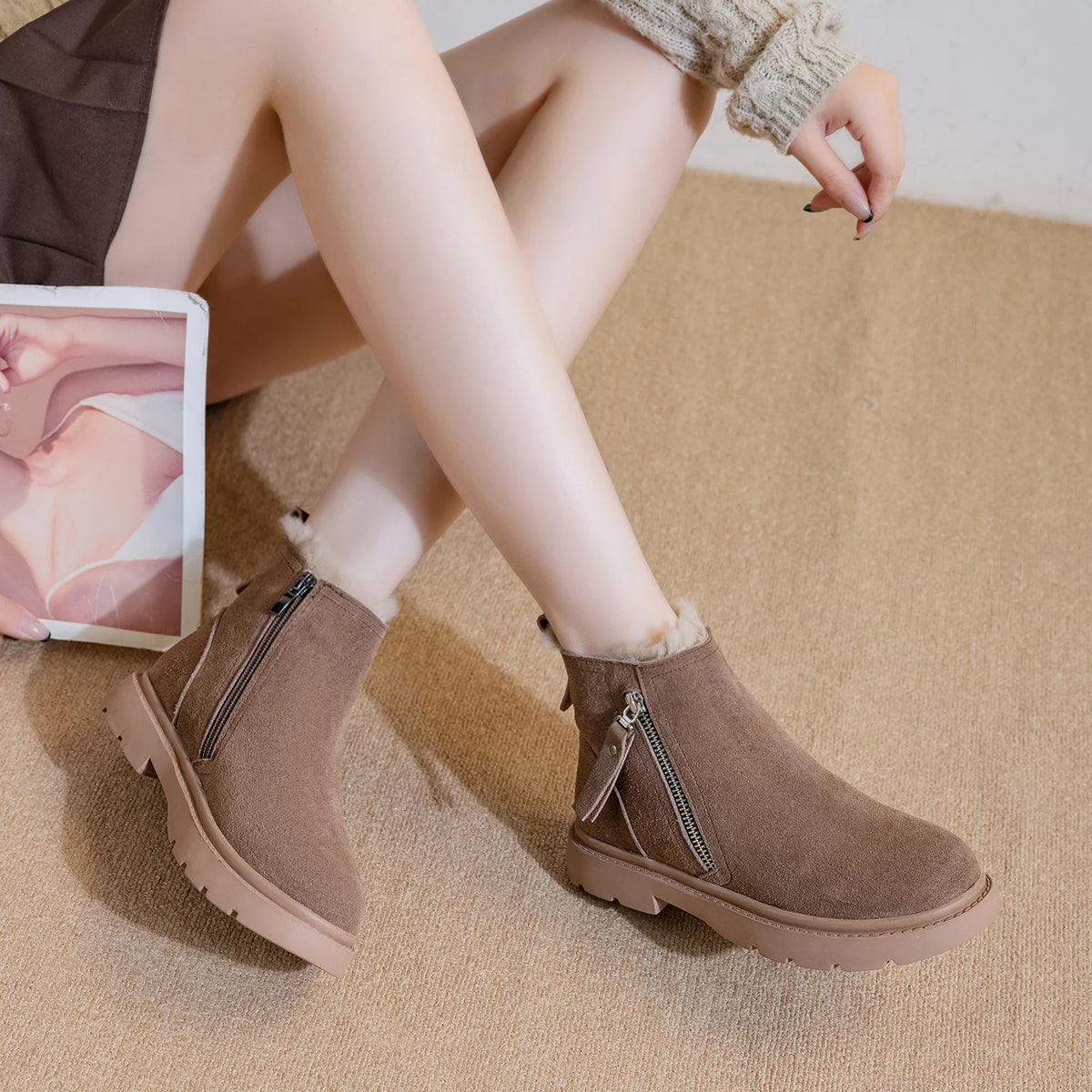 Modelo sentada usando botines de ante marrones con cremallera lateral y forro de felpa, sosteniendo una revista de moda