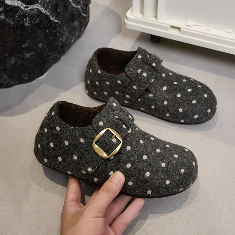 Zapatillas planas grises oscuras con lunares blancos y hebilla dorada junto a piedra negra