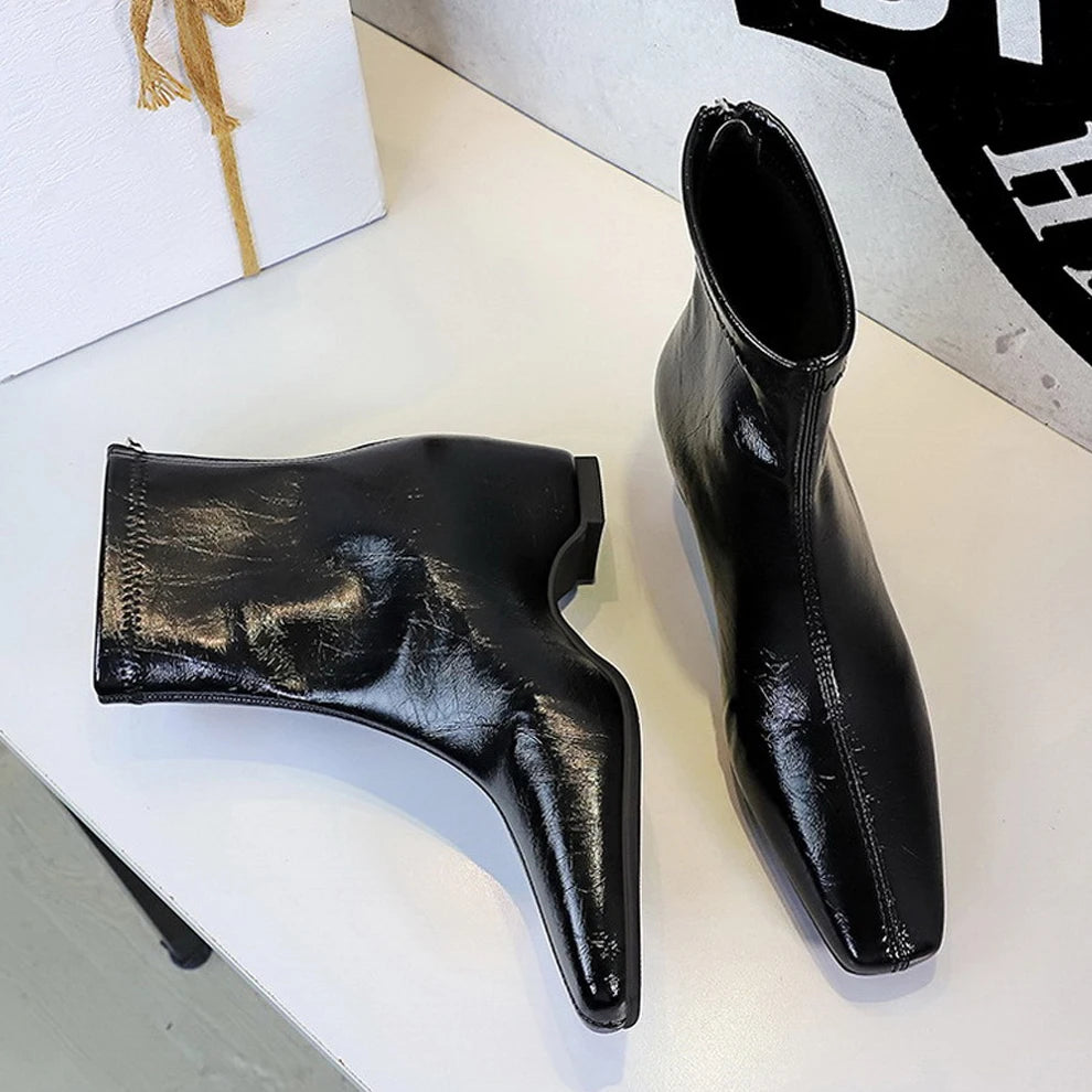 Botas pretas brilhantes de bico quadrado com painel elástico ao lado de uma caixa de presente na mesa