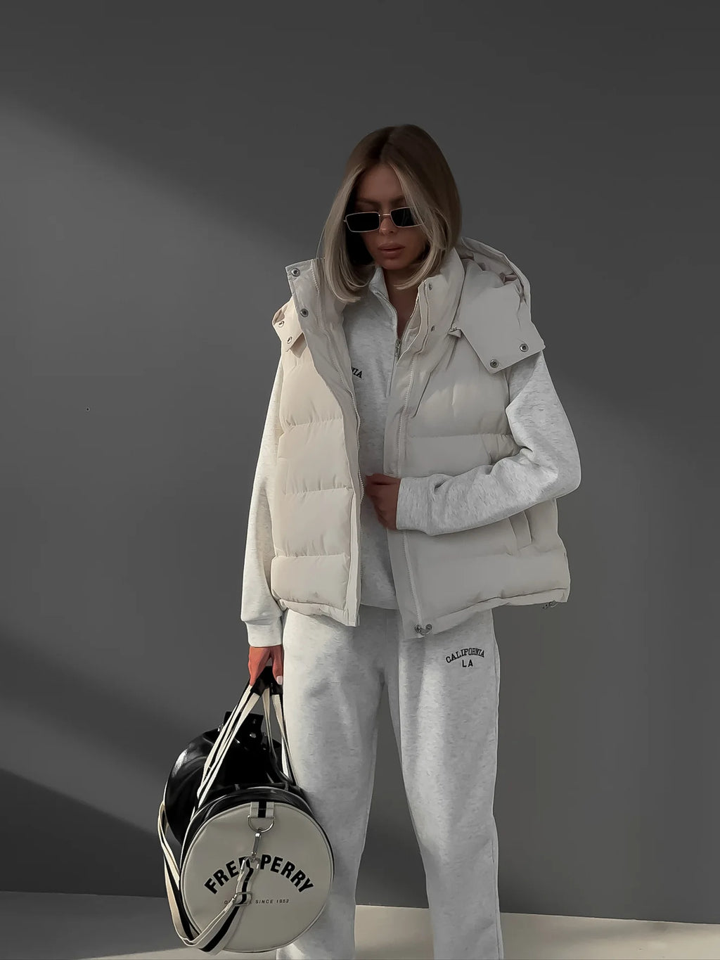 Foto corporal inteira de modelo com colete acolchoado branco com capuz, segurando mala redonda