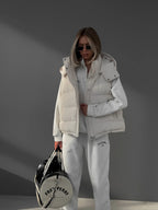 Foto corporal inteira de modelo com colete acolchoado branco com capuz, segurando mala redonda