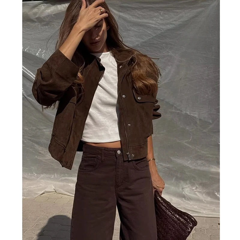 Modelo usa jaqueta cropped castanha-escura aberta com fecho e botões de pressão sobre t-shirt branca e calças.
