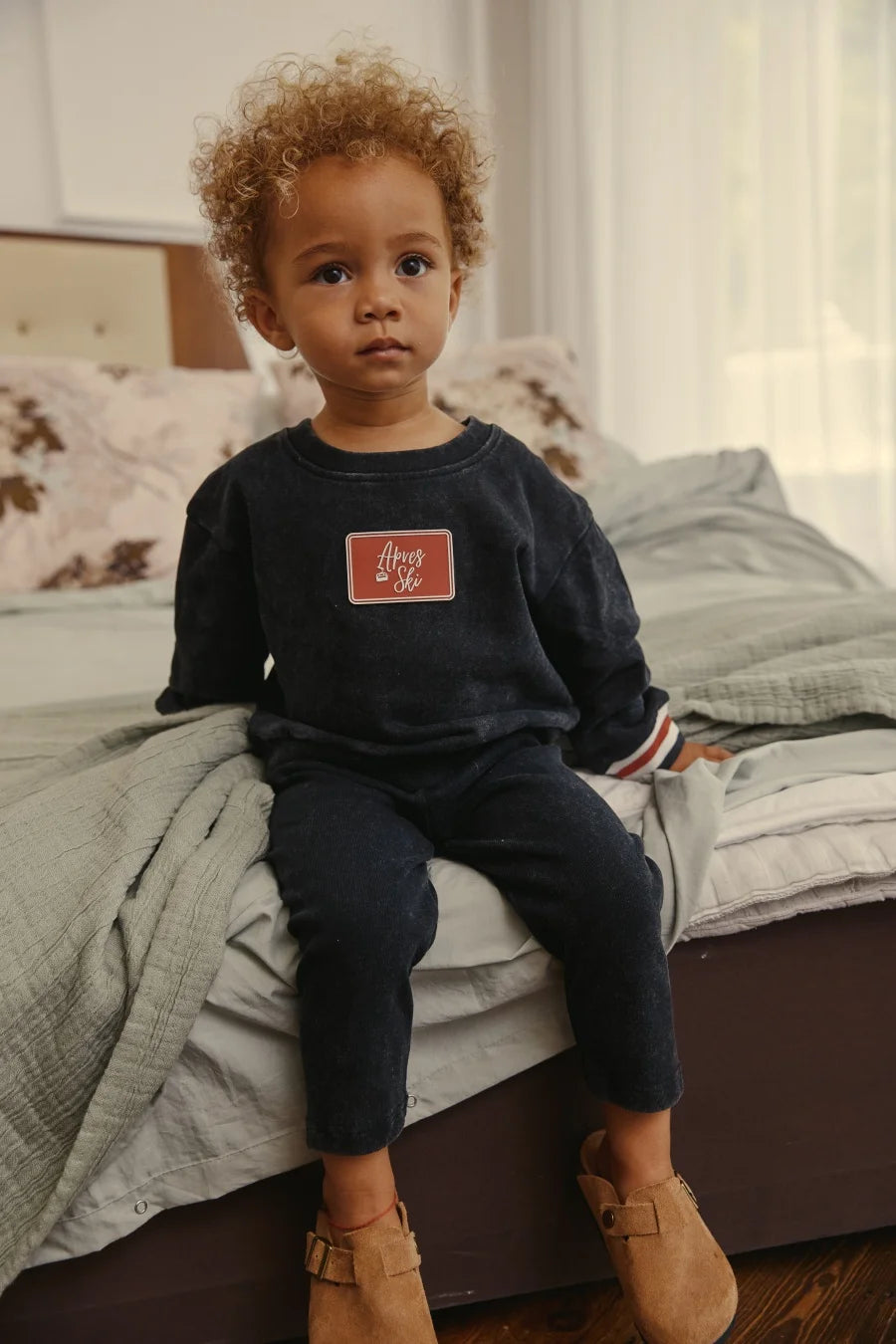 Criança pequena na cama com suéter preto lavado e joggers com remendo vermelho proeminente Apres Ski