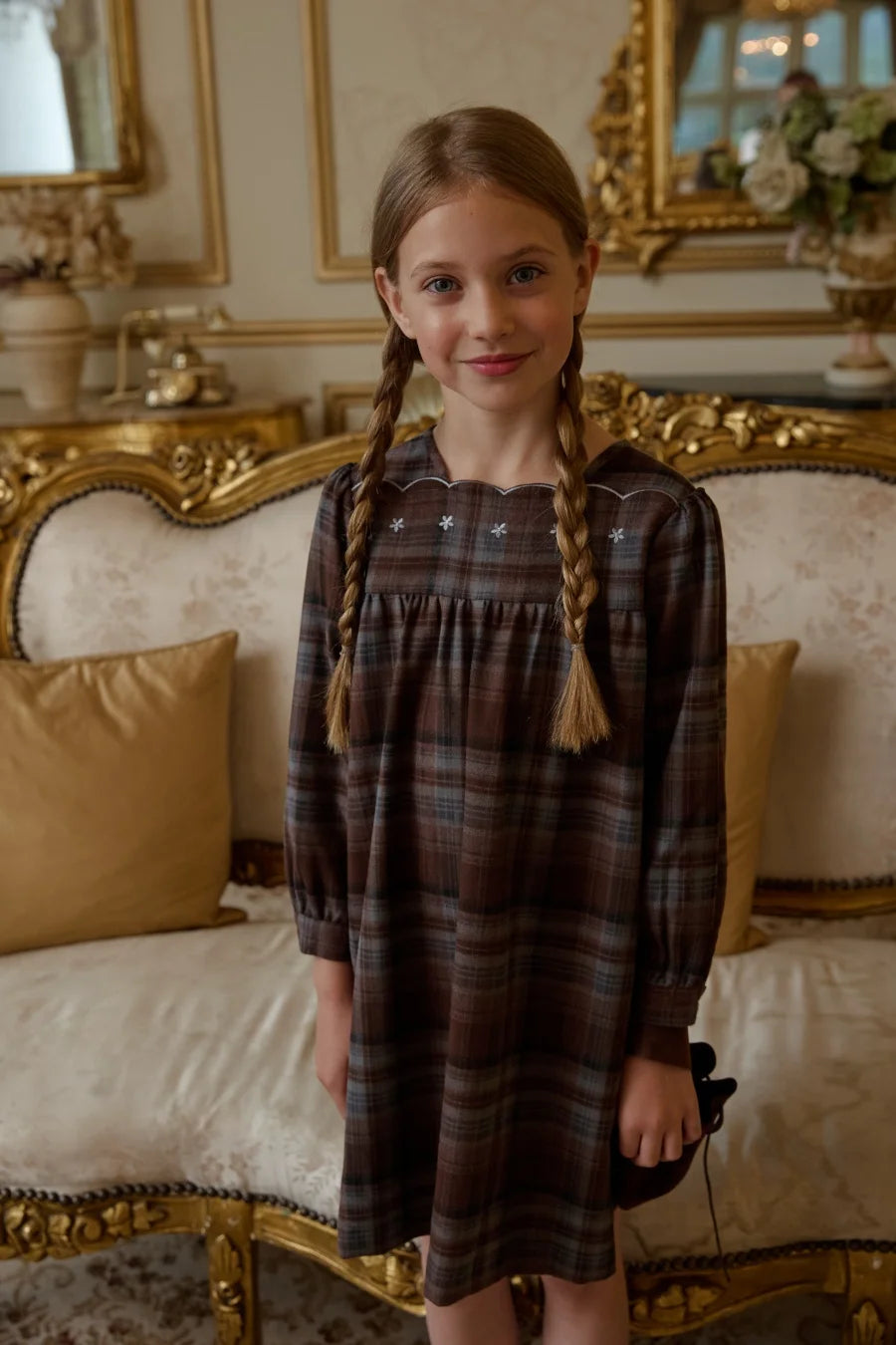 Menina em vestido de xadrez castanho com detalhes bordados brancos, num elegante quarto com espelhos dourados.