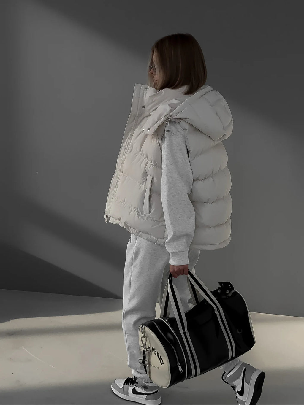 Colete acolchoado branco com capuz e estofamento horizontal, usado sobre conjunto desportivo cinza