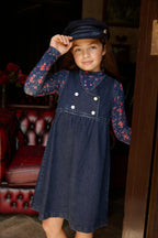 Menina com vestido jumper de ganga azul-marinho, painel assimétrico e botões brancos sobre uma gola alta floral e boné.