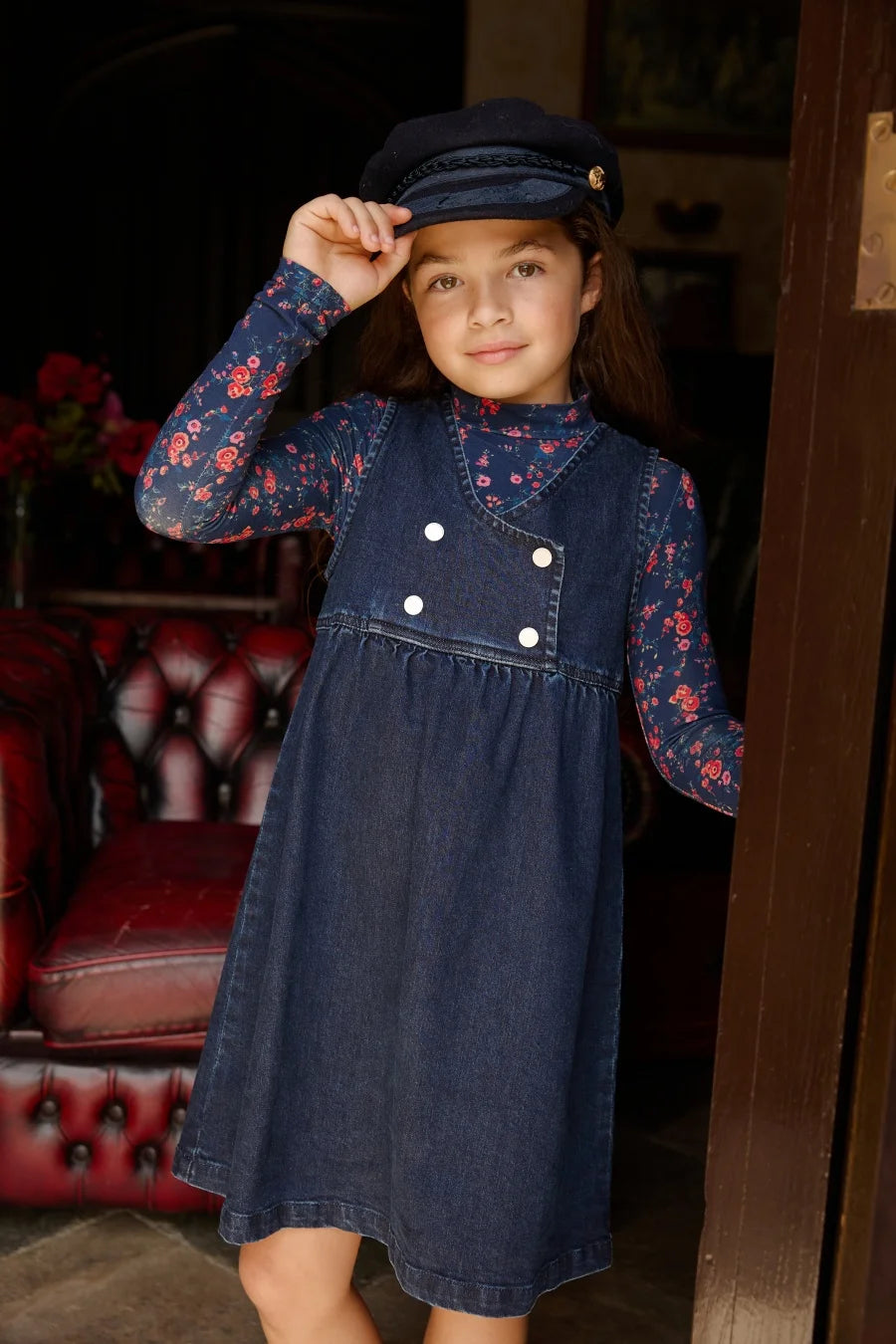 Menina com vestido jumper de ganga azul-marinho, painel assimétrico e botões brancos sobre uma gola alta floral e boné.