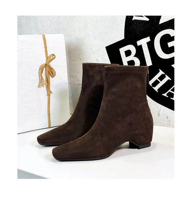 Dunkelbraune Wildlederschnürsenkel-Stiefel mit quadratischer Spitze und niedrigem Blockabsatz auf weißer Displayfläche neben Geschenkbox.