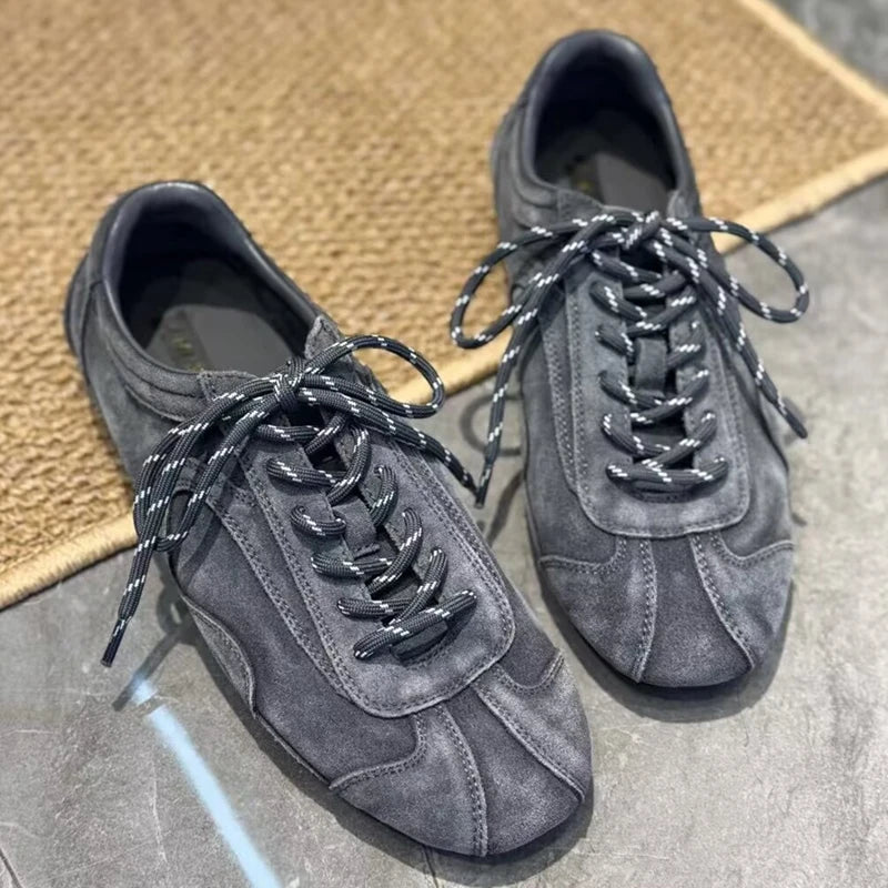 Dark grey suede sneakers with speckled laces on beige woven mat against stone background