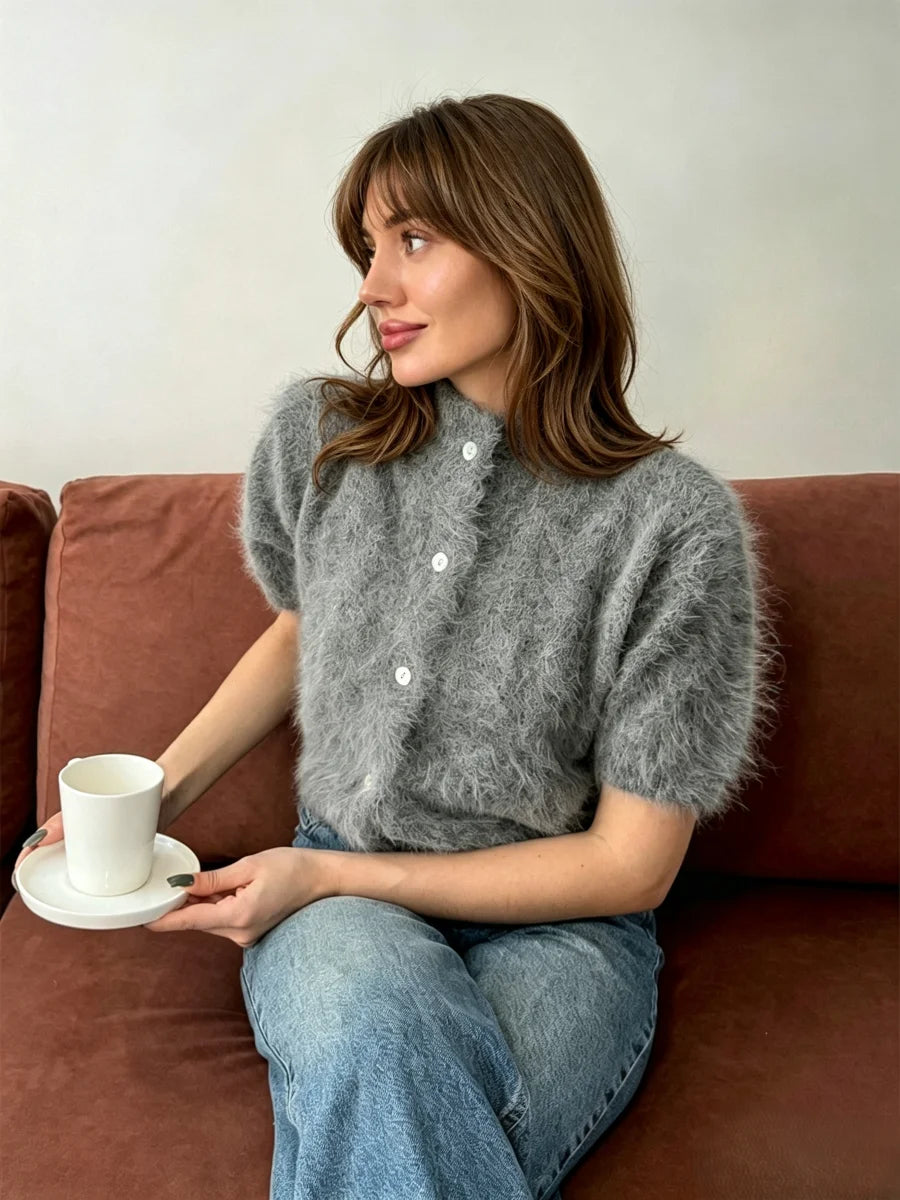 Mulher sentada em sofá cor ferrugem com cardigan curto de pelúcia cinza com botões brancos, segurando chávena de café e usando acessórios