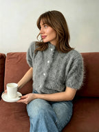 Mulher sentada em sofá cor ferrugem com cardigan curto de pelúcia cinza com botões brancos, segurando chávena de café e usando acessórios