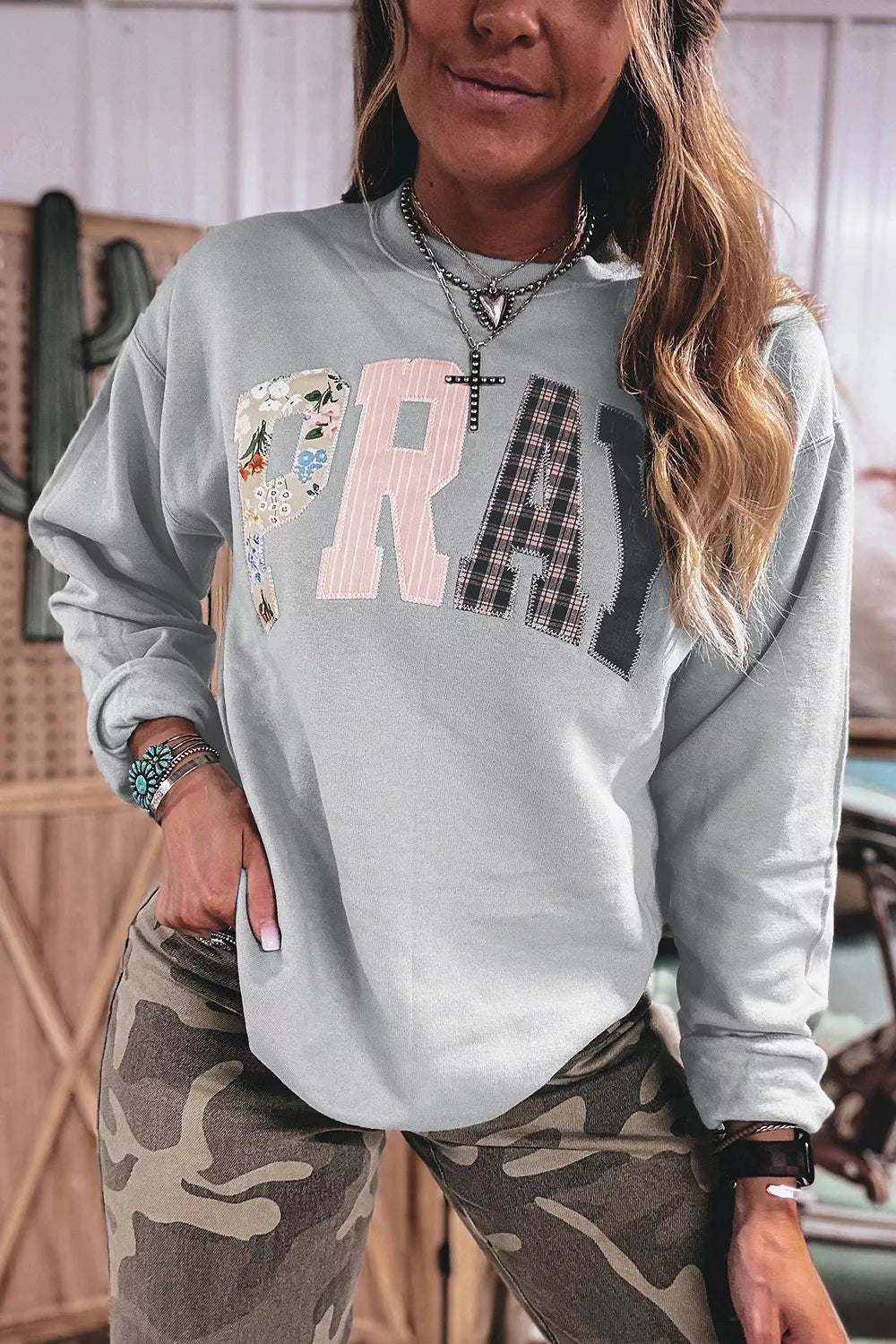Model in light grey sweatshirt with mixed-media PRAY letters: floral P, pink striped R, plaid A, dark Y. Styled with camo