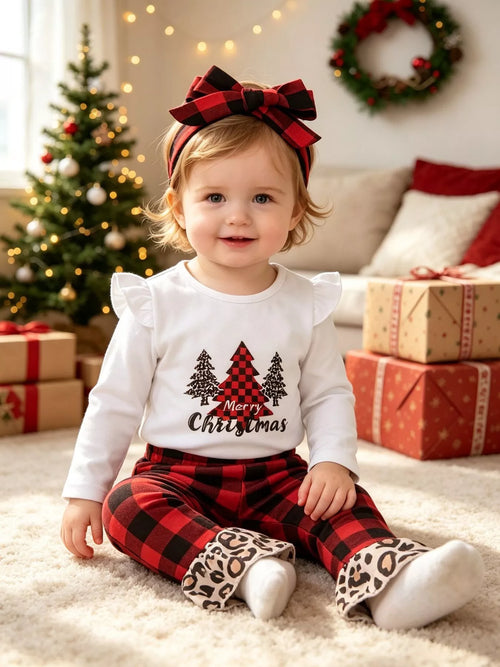 Smiling toddler on white rug wearing festive plaid outfit with tree graphics and large bow headband near gifts
