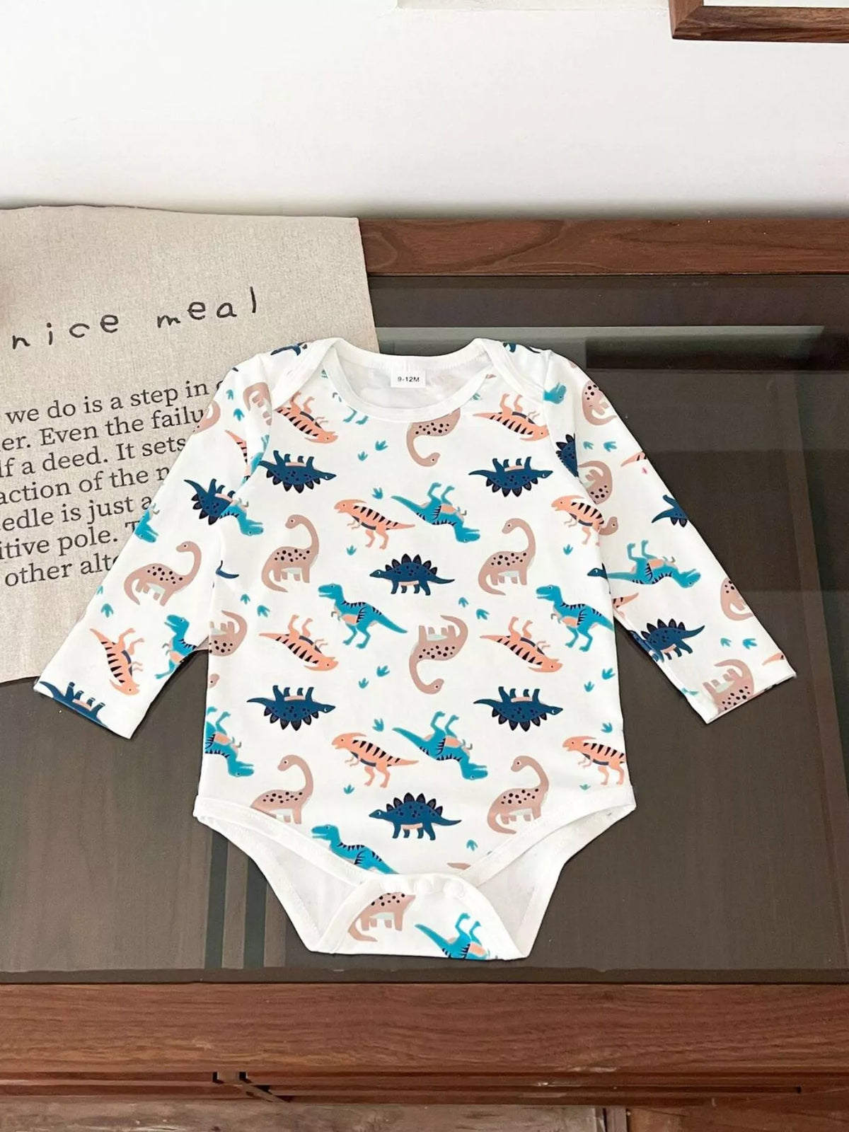 White long-sleeve bodysuit with colorful multi-dinosaur print pattern laid flat on dark wooden surface