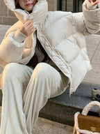 Seated model in cream puffer jacket adjusting high collar while sitting on stone steps next to bag