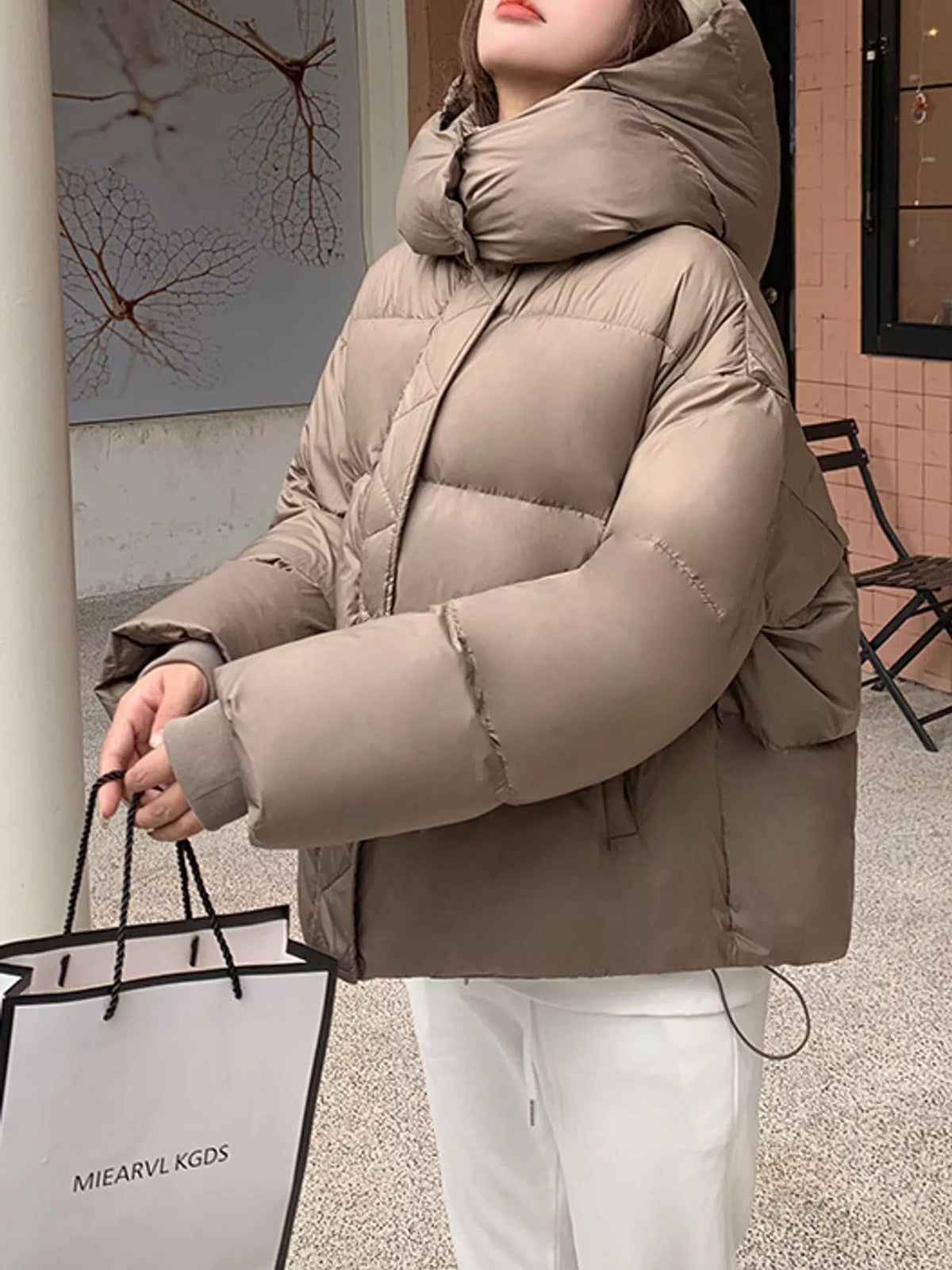 Model wearing taupe quilted hooded puffer jacket outdoors, holding shopping bag with urban styling